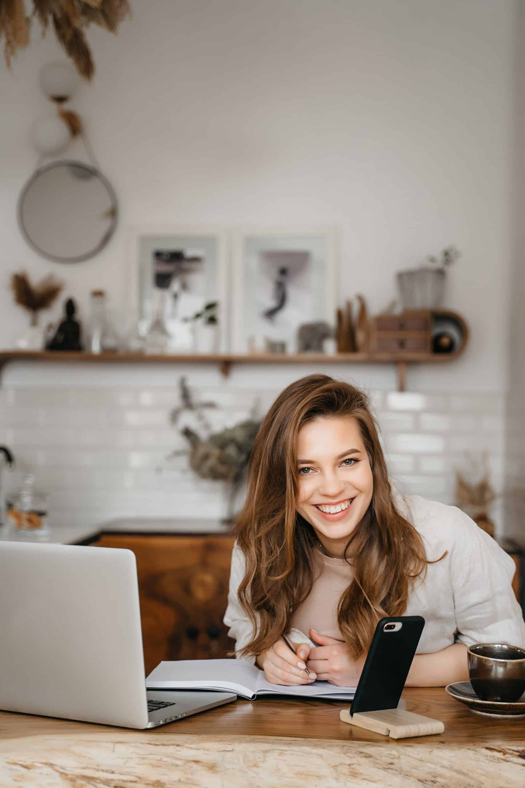 Happy attractive young woman sitting in her kitchen at the counter with a laptop smiling at the camera. High quality photo